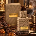 French Avenue Sacred Smoke — Kotak dan botol emas bertekstur seperti reben, dikelilingi serpihan kayu oud dan ketulan resin amber, dengan asap naik dari pembakar hiasan di latar belakang.