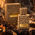 French Avenue Sacred Smoke — Kotak dan botol emas bertekstur anyaman, diletakkan di atas batu gelap dengan kristal ambar dan kayu oud, asap naik.