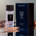 Bujairami Shadow Script - Bujairami 100ml Extrait, clear faceted glass bottle with dark cap, next to navy blue presentation box with silver logo, on reflective surface with dried wheat and seed pods.