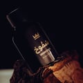 Gulf Orchid Shumookh Parfum 100ml by Gulf Orchid — A dark, elegant perfume bottle with gold Arabic script and a crown logo, resting on textured, reddish-brown wood against a dark background. | Gulf Orchid Shumookh Parfum 100ml by Gulf Orchid — Botol minyak wangi gelap yang elegan dengan tulisan Arab emas dan logo