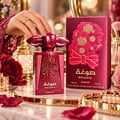 Asdaaf Sougha by Asdaaf 100ml EDP Women — A woman's hand with a ring is touching the red perfume bottle's decorative cap. The bottle and its red box with floral design are on a marble surface with roses and jewelry.
