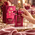 Asdaaf Sougha 100ml EDP Women — Red perfume bottle with gold speckles and ornate cap, next to a red gift box with a floral cutout design and bow, surrounded by pink peonies and gold accents.