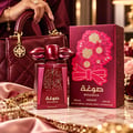 Asdaaf Sougha by Asdaaf 100ml EDP Women — A woman's hand with a ring is touching the red perfume bottle's decorative cap. The bottle and its red box with floral design are on a marble surface with roses and jewelry.
