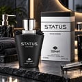 TADangel Status by TADangel 100ml EDP Men — Botol minyak wangi hitam matte dengan penutup perak dan reben hiasan, diletakkan di atas permukaan gelap bertekstur bersebelahan kotak perak dan hitamnya. Tuala kelabu gelap berlipat kelihatan di sebelah kiri.