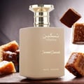 Paris Corner Taskeen Caramel Cascade 100ml — A beige perfume bottle with a gold cap sits on a dark surface, surrounded by pieces of caramel candy dusted with powdered sugar. | Paris Corner Taskeen Caramel Cascade 100ml — Botol minyak wangi berwarna krim dengan penutup emas duduk di atas permukaan gelap, dikelilingi