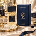 Bujairami Tempted 100ml Extrait Perfume Unisex — Clear faceted glass bottle with gold accents and black cap, placed on a gold tray next to its navy blue presentation box. White flowers and marble accents in the background.