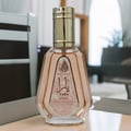 Ard Al Zaafaran Yara Eau De Parfum 50ml — A clear glass perfume bottle with a pale pink liquid and a gold cap, featuring a decorative label with Arabic script and floral motifs, set against a blurred background of furniture. | Ard Al Zaafaran Yara Eau De Parfum 50ml — Botol minyak wangi kaca jernih dengan cecair merah