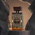 Zimaya Amber Is Great Eau De Parfum 100ml — A dark glass perfume bottle with gold art deco detailing and amber liquid sits on a stone pedestal, framed by rough, light-colored rock formations against a warm gradient background. | Zimaya Amber Is Great Eau De Parfum 100ml — Botol minyak wangi kaca gelap dengan perincian