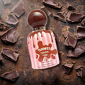 Grandeur Tubbees Chocolate Fudge by Grandeur 50ml EDP — A pink and white striped perfume bottle with chocolate drizzle design, topped with a chocolate-colored sphere cap, surrounded by broken pieces of dark chocolate on a textured brown surface. | Grandeur Tubbees Chocolate Fudge by Grandeur 50ml EDP — Botol minyak