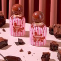 Tubbees Chocolate Fudge by Grandeur 50ml EDP — Two bottles of chocolate fudge-scented perfume with pink and white striped labels, surrounded by chocolate brownies and chocolate shards on a light pink surface with a pink curtain backdrop. | Tubbees Chocolate Fudge by Grandeur 50ml EDP — Dua botol minyak wangi beraroma