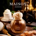 Maison Asrar Vanilla Voyage — Botol minyak wangi bulat keemasan dengan penutup bertekstur warna krim diletakkan di atas permukaan gelap. Latar belakang kelihatan kamera vintaj kabur dan pencuci mulut putih.