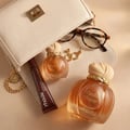 Maison Asrar Vanilla Voyage by Maison Asrar 100ml EDP Unisex — Dua botol minyak wangi kaca berpenutup berjalur emas dan perincian emas, tiub pewarna bibir coklat, anting-anting bulat emas, dan cermin mata kerangka kura-kura tersusun di atas beg tangan krim.